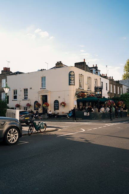 A charming corner building on Chiswick High Road, featuring a white exterior with decorative hanging flower baskets and window boxes filled with colorful blooms. The ground floor appears to house a pu