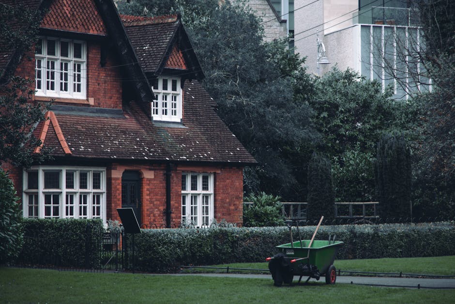 A red-brick house with a steep, tiled roof features multiple white-framed windows, some with small panes, surrounded by a neatly maintained garden. In the foreground, a lush, green lawn is bordered by